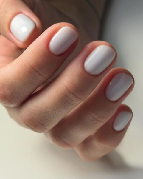Zdjęcie ze zbliżeniem na krótkie, owalne paznokcie pomalowane lakierem w odcieniu milky white. Dłoń pozuje oparta o biały blat.