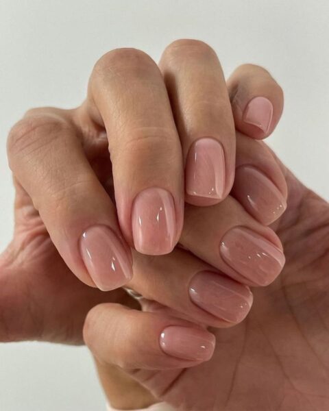 Zdjęcie dłoni ze zbliżeniem na krótkie, beżowe paznokcie z mocnym połyskiem. Dłonie pozują tak, by prezentować manicure.