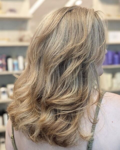 Zdjęcie dojrzałej kobiety w salonie fryzjerskim. Pozuje tyłem i prezentuje uczesanie: cieniowane włosy do ramion i z blond pasemkami w odcieniu ash blonde.