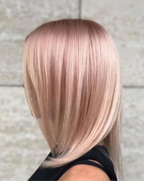 Zdjęcie modelki stojącej bokiem, prezentującej fryzurę typu bob w kolorze blond w pastelowym, różowym podtonem. Ubrana jest w czarny top na ramiączkach.