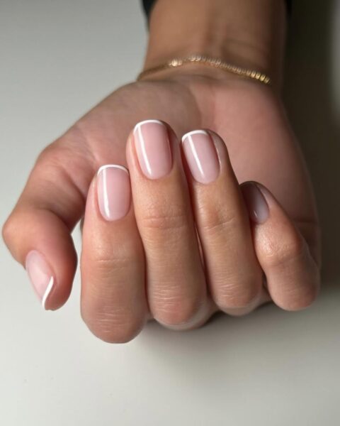 Zdjęcie dłoni ze zbliżeniem na krótkie, kwadratowe paznokcie ze stylizacją micro french manicure. Dłoń pozuje na blacie białego stołu.