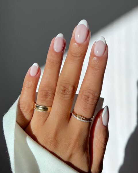 Zdjęcie dłoni ze zbliżeniem na długie, owalne paznokcie z rounded french manicure. Na palcach znajdują się złote pierścionki.
