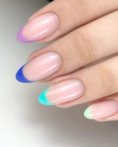 Zdjęcie dłoni ze zbliżeniem na długie, migdałowe paznokcie ze stylizacją french manicure z kolorowymi końcówkami. Dłoń pozuje na białym blacie stołu.