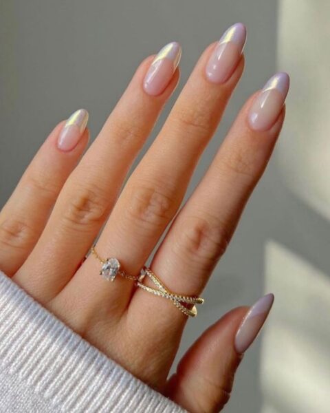Zdjęcie dłoni z długimi, migdałowymi paznokciami pomalowanymi na wzór french manicure z fioletową, perłową końcówką Na palcach znajdują się dwa złote pierścionki.