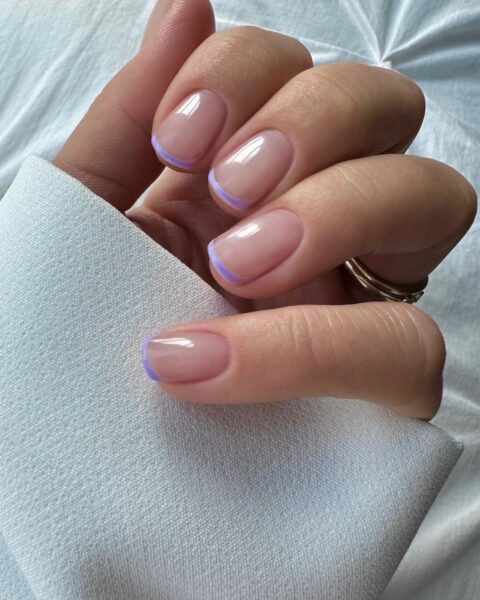 Zdjęcie dłoni z krótkimi, kwadratowymi paznokciami w stylu french manicure w kolorze fioletowym, zamiast białym. Dłoń pozuje na tle białego materiału.