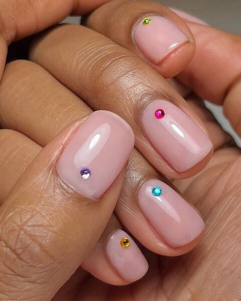 Zdjęcie dłoni ze zbliżeniem na manicure z krótkimi, kwadratowymi paznokciami pomalowanymi na kolor pink nude oraz ozdobione kolorowymi, kryształkami umieszczonymi po jednym na każdym paznokciu.