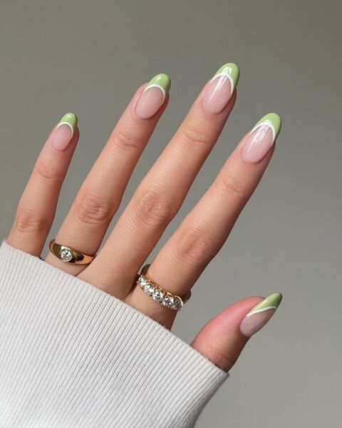 Zdjęcie dłoni ze zbliżeniem na długie, migdałowe paznokcie ozdobione wzorem french manicure z podwójną linią przy brzegu paznokci: pistacjową i białą. Na palcach znajdują się dwa złote pierścionki.