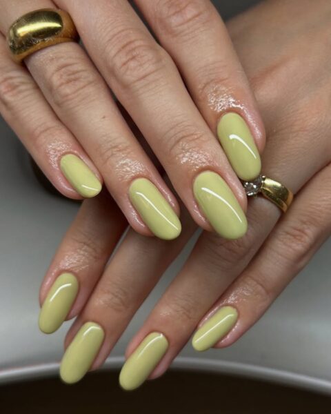 Zdjęcie dłoni z długimi, migdałowymi paznokciami w kolorze pistacjowym. Dłonie układają się tak, by prezentować manicure. Widać na palcach złote pierścionki.