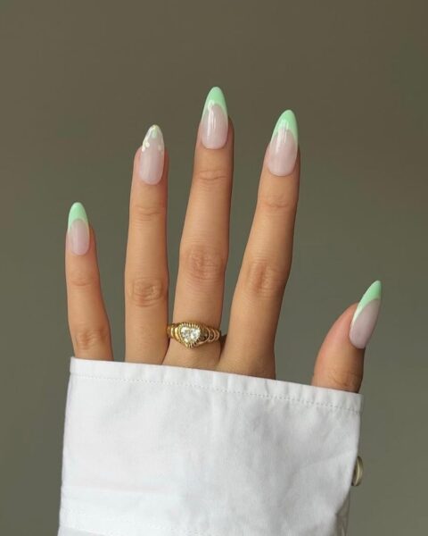 Zdjęcie dłoni z długimi, migdałowymi paznokciami ze zdobieniem french manicure w kolorze miętowym. Paznokieć palca serdecznego jest ozdobiony motywem kwiatków. Na palcu środkowym znajduje się złoty pierścionek.
