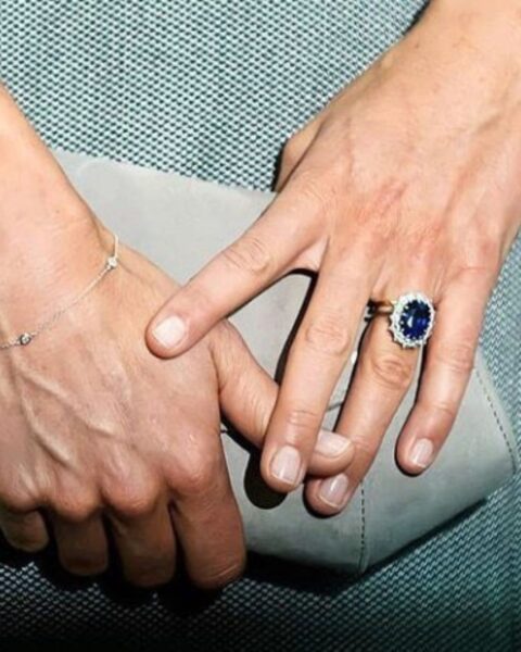 Zdjęcie ze zbliżeniem na dłonie Kate Middleton, która nosi paznokcie princess nails, czyli krótkie i w kolorze nude. Na palcu widać pierścionek z niebieskim kamieniem. Trzyma torebkę typu kopertówkę w szałwiowym kolorze, którą widać na tle zielonej torebki.