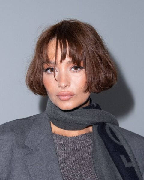 Na zdjęciu znajduje się celebrytka Taylor Lashae, która ma fryzurę typu riviera bob. Co to za fryzura? Lekko falowana, podkręcona, z grzywką. Ubrana jest w szary sweter, szarą marynarkę oraz szary szalik.