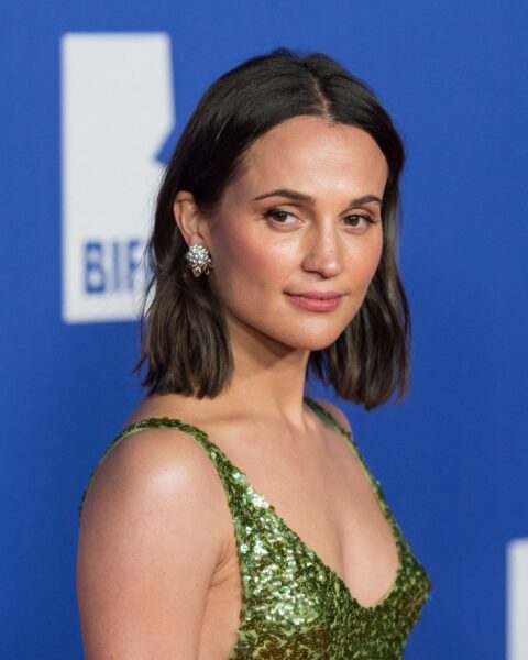 Na zdjęciu znajduje się aktorka Alicia Vikander, która pozuje na niebieskiej ściance. Ma fryzurę typu bob, ciemną, lekko falowaną. Ubrana jest w cekinową sukienkę na ramiączkach. W uszach nosi srebrne kolczyki.