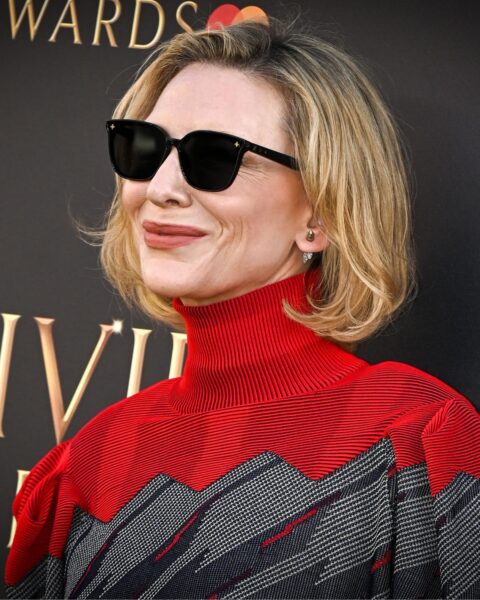 Zdjęcie aktorki Cate Blanchett ubranej w czarno-szaro-czerwony sweter z golfem. Na nosie ma okulary przeciwsłoneczne. Ma krótkie, blond włosy na boba. Pozuje na ściance.