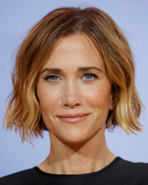 Zdjęcie aktorki Kristen Wiig. To portret ze zbliżeniem na jej krótkie, brązowe włosy w stylu boba. Ubrana jest w czarną bluzkę bez rękawów.