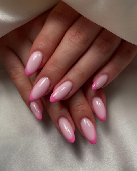 Zdjęcie dłoni ze zbliżeniem na długie, migdałowe paznokcie. Są pomalowane w stylu french manicure z różowymi kreskami na brzegach paznokci. Dłonie pozują tak, by prezentować manicure, w otoczeniu białego materiału.