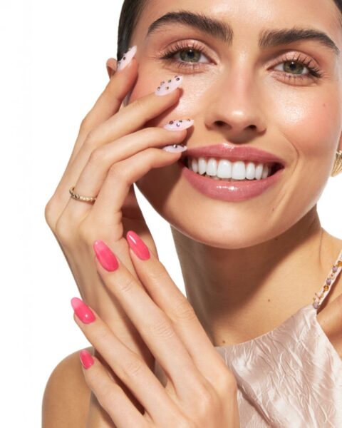 Zdjęcie kobiety w ciemnych, długich włosach. Przykłada do twarzy dłonie i prezentuje manicure: na jednej ręce nude z kryształkami, a na drugiej różowe z efektem jelly.