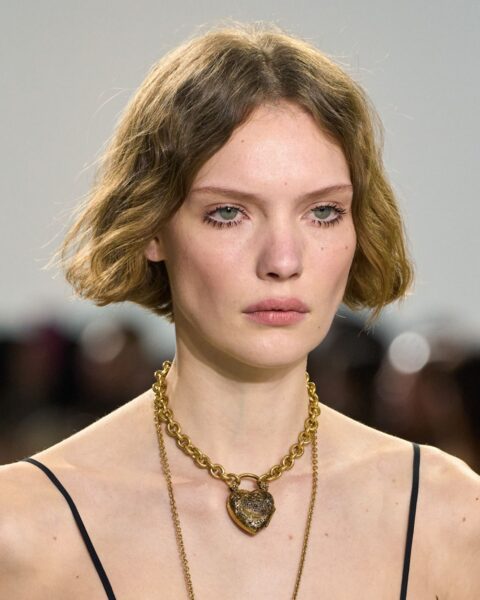 Zdjęcie potretowe modelki na pokazie marki Chloé. Ma krótkie włosy do brody, jak krótki bob z kolorem tobacco bronde. Ubrana jest w koronkowy top na ramiączkach. Na szyi ma masywny, złoty naszyjnik i drugi, długi, wiszący. Ma mocno wytuszowane rzęsy.