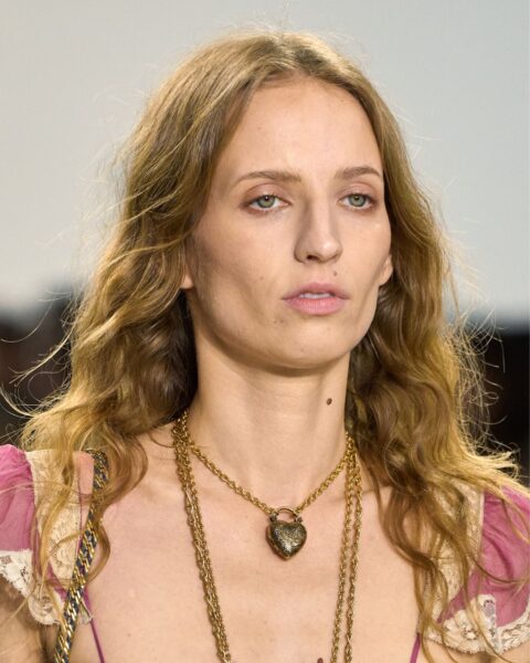 Zdjęcie portretowe modelki na wybiegu podczas prezentacji kolekcji marki Chloé. Kobieta ma długie, kręcone brązowo-blond włosy. Ubrana jest w różową sukienkę z koronkowym wykończeniem. Na szyi ma naszyjnik z zawieszką w kształcie serca, oraz długi, wiszący naszyjnik.