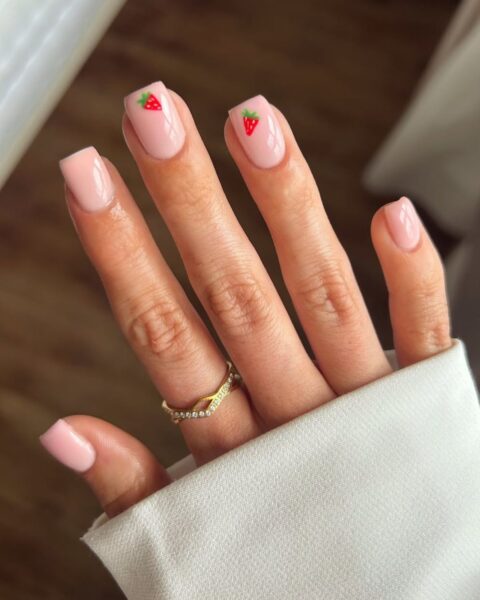 Zdjęcie dłoni ze zbliżeniem na długie, kwadratowe paznokcie w kolorze pink nude, a na dwóch paznokciach jest namalowany wzór truskawki. Na palcu wskazującym znajduje się złoty pierścionek. Dłoń pozuje na tle podłogi.