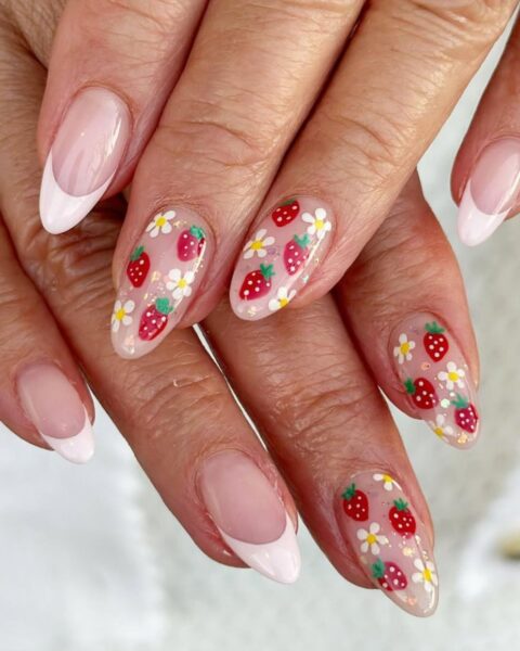 Zdjęcie dłoni z długimi, migdałowymi paznokciami. Są pomalowane w stylu french manicure i do tego ozdobione wzorem truskawek i kwiatków. Dłonie pozują tak, by prezentować manicure.