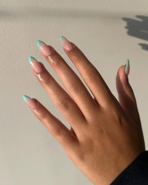 Zdjęcie dłoni, które mają długie, owalne paznokcie pomalowane w stylu french manicure z turkusową końcówką. Dłoń układa się na tle ściany tak, by prezentować manicure.