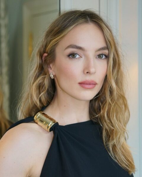 Zdjęcie aktorki Jodie Cormer, która ma modne włosy na lato 2025. Trendy stawiają na jasne blondy, a ona ma najmodniejszy: słoneczny blond. Ma falowane włosy do ramion. Ubrana jest w czarny top halter ze złotą aplikacją.