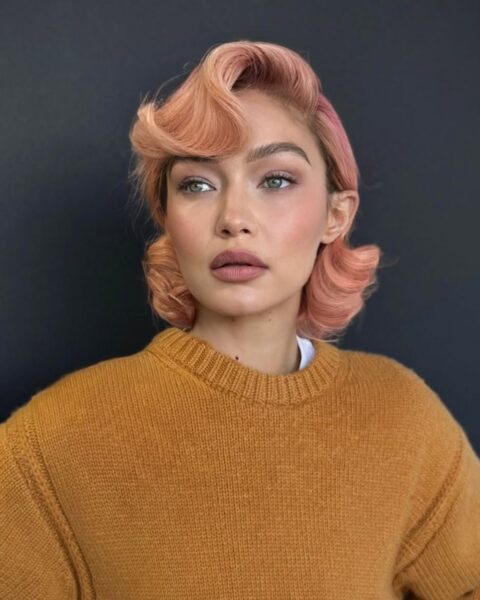 Zdjęcie modelki Gigi Hadid, która ma modny, morelowy blond na lato 2025. Włosy do ramion, podkręcone w stylu hollywoodzkim. Ubrana jest w musztardowy sweter.