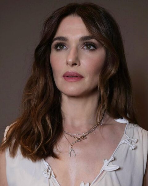 Zdjęcie aktorki Rachel Weisz, która ma ciemne włosy do ramion w odcieniu czekoladowego brązu. Ubrana jest w lekką, kremową bluzkę bez rękawów wykończoną aplikacją w kształcie listków. Na szyi ma kilka naszyjników. Patrzy w dal.