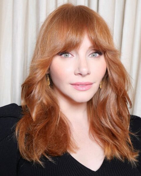 Zdjęcie Bryce Howard w długich, rudych włosach, która ma modną grzywkę wispy fringe. Ubrana jest w czarną bluzkę z bufkami i pozuje na tle kremowej zasłony.
