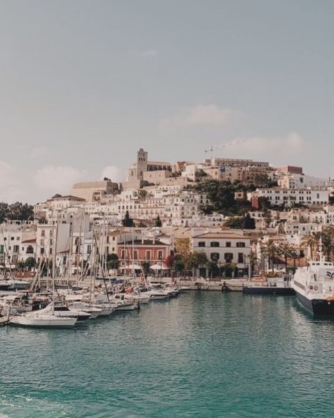 Zdjęcie przedstawiające panoramiczny widok na stare miasto Ibiza Eivissa od strony portu. Widać morze, port, statki, budynki, a także wzgórze z wysokim zamkiem.
