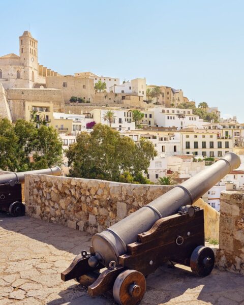 Zdjęcie przedstawiające widok na stare miasto w Eivissa na Ibizie z perspektywy tarasu. Widać zamek z ratuszem i wieżą, domostwa, a na pierwszym planie armatę strzelniczą opartą o starożytny murek.