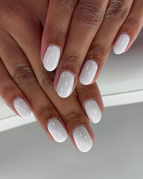 Zdjęcie dłoni z krótkimi, owalnymi paznokciami w kolorze białym z mocnym połyskiem w stylu glossy white manicure. Dłonie pozują ułożone jedna na drugą w taki sposób, by prezentować manicure.
