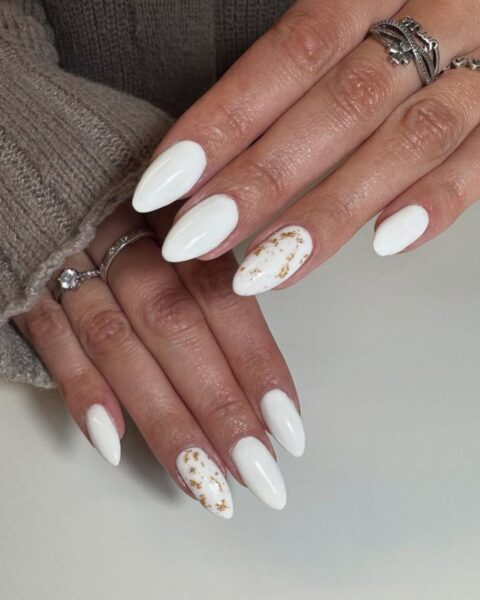 Zdjęcie dłoni z długimi, migdałowymi paznokciami. Mają biały manicure, a na paznokciach palców serdecznych znajdują się drobne, złote zdobienia. Dłonie pozują na białym blacie, prezentując manicure.