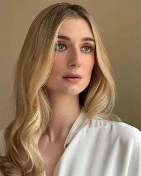 Zdjęcie aktorki Elisabeth Debicki, która ma elegancki blond dla kobiet z klasą: długie, złote blond włosy. Ubrana jest w białą koszulę i patrzy na tym zdjęciu gdzieś w dal.