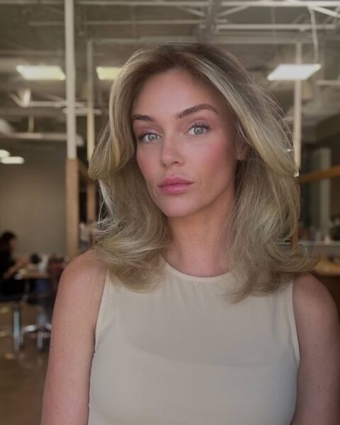 Zdjęcie kobiety z blond pasemkami na półdługich włosach do ramion. Ubrana jest w beżowy top bez rękawów. Pozuje w dużym salonie fryzjerskim.