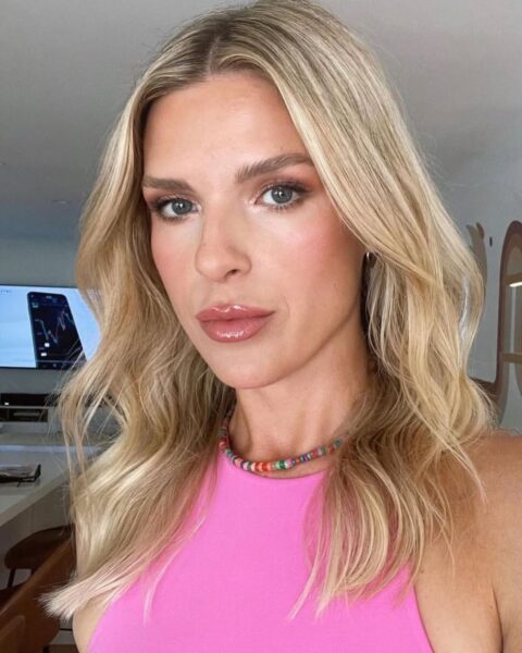 Zdjęcie kobiety z blond pasemkami na półdługich włosach do ramion. Ubrana jest w różowy top bez rękawów. Ma na szyi naszyjnik z kolorowych koralików. Pozuje w salonie fryzjerskim.