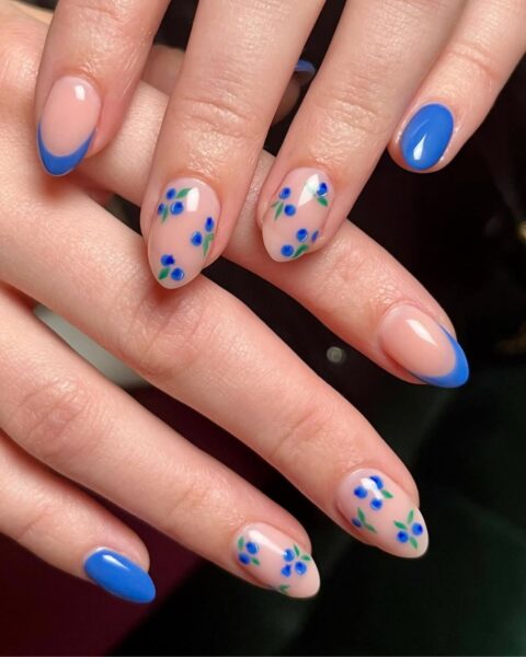 Zdjęcie dłoni ze zbliżeniem na długie, migdałowe paznokcie ozdobione niebieskim french manicure oraz wzorem borówek.