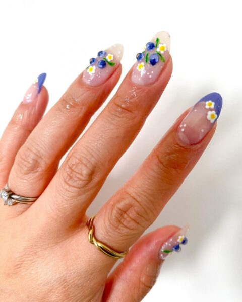 Zdjęcie dłoni z długimi, migdałowymi paznokciami pomalowanymi na wzór french manicure w kolorze białym i niebieski oraz wzorem borówek wykonanych strukturalnym wzorem. Na palcach widać złote pierścionki.