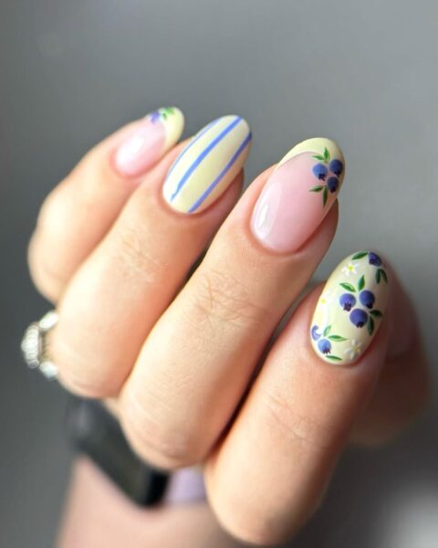 Zdjęcie dłoni ze zbliżeniem na długie, migdałowe paznokcie. Są ozdobione wzorem pasków, french manicure, kwiatów i borówek w kolorze żółtym i niebieskim.