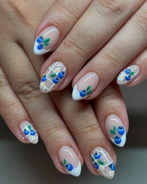 Zdjęcie dłoni z długimi, migdałowymi paznokciami z białym french manicure. Są dodatkowo ozdobione wzorem borówek i kratki. Dłonie układają się tak, by prezentować manicure.