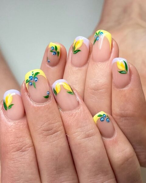 Zdjęcie dłoni ze zbliżeniem na długie, kwadratowe paznokcie ze wzorem french manicure w kolorze żółto-białym i ozdobione wzorem borówek oraz cytrynek.