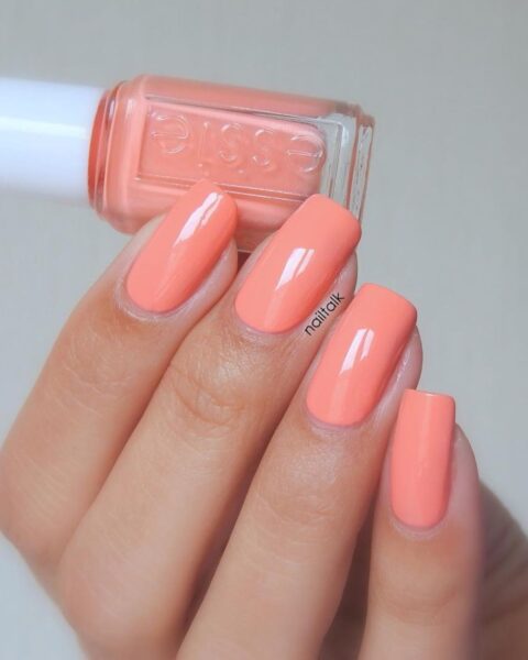 Zdjęcie dłoni ze zbliżeniem na brzoskwiniowy manicure, czyli długie, kwadratowe paznokcie w kolorze brzoskwiniowym. Dłoń trzyma buteleczkę lakieru marki Essie.