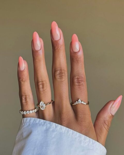Zdjęcie dłoni z długimi, migdałowymi paznokciami z brzoskwiniowym ombre manicure. Na palcach znajduje się kilka pierścionków. Widać mankiet białej koszuli.
