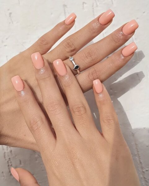 Zdjęcie dłoni z kwadratowymi paznokciami pomalowanymi lakierem w kolorze rozbielonej brzoskwini. Dłonie pozują tak, by prezentować manicure. Na środkowym palcu jednej z dłoni widać srebrny pierścionek z dwoma kamieniami.