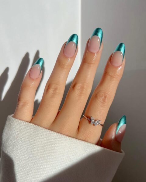 Zdjęcie dłoni z długimi, owalnymi paznokciami z french manicure, ale zamiast białej końcówki są zielone, chromowane zdobienia. Na palcu wskazującym widać delikatny, srebrny pierścionek z motywem motylka i cyrkoniami.
