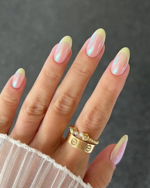 Zdjęcie dłoni z długimi, owalnymi paznokciami pomalowanymi na wzór french manicure z żółtą końcówką oraz lśniącym wykończeniem chromu. Na palcu wskazującym znajdują się dwa złote pierścionki Widać fragment siateczkowej, białej bluzki.