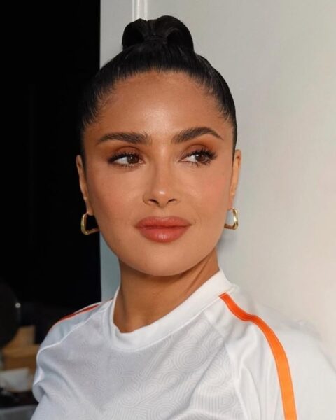 Zdjęcie aktorki Salmy Hayek. Wygląda pięknie, bo zastosowała dojrzałe triki makijażowe i pomalowała oczy matowym cieniem. Ma ciemne włosy związane gładko w kok. Ubrana jest w białą bluzkę z pomarańczowymi lampasami. W uszach ma złote kolczyki.
