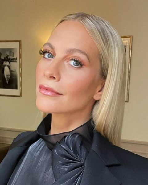 Zdjęcie celebrytki Poppy Delevingne, która ma makijaż dla dojrzałej cery z pomalowanymi tylko górnymi rzęsami. Ma blond, długie, proste włosy rozpuszczone na plecy. Ubrana jest w czarną bluzkę ze stójką. Pozuje w pokoju hotelowym, za nią widać ścianę z obrazami.