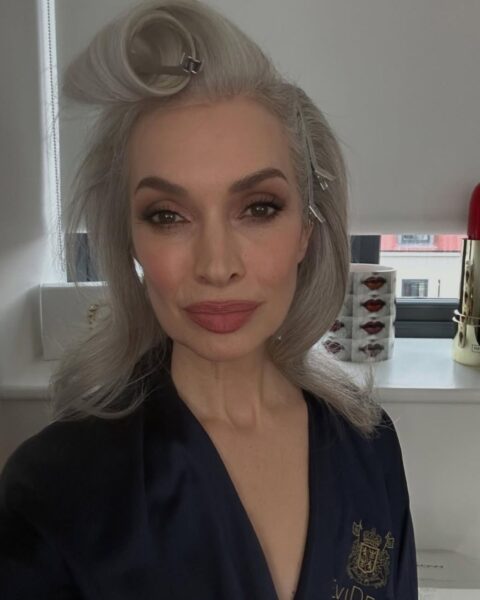 Zdjęcie modelki Kelly Brennan. Ma długie, siwe włosy podkręcone na wałki. Ma tez elegancki makijaż dla dojrzałej cery z podkreślonymi oczami, ustami oraz konturowaniem różem. Ubrana jest w granatowy szlafrok z haftem.