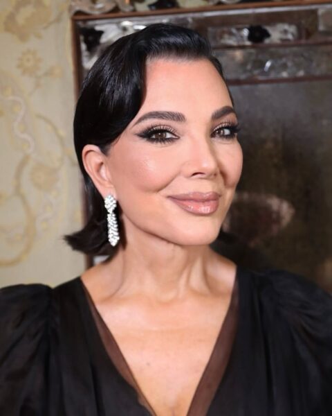 Zdjęcie portretowe celebrytki Chirs Jenner. Ma ciemne włosy do ramion, gładko zaczesane na bok, lekko uniesione w stylu retro, lśniące. Ubrana jest w wieczorową, czarna suknię z dekoltem V i bufkami. W uszach ma lśniące, wiszące kolczyki z brylantami. Ma też mocny makijaż oczu oraz modnie pomalowane usta konturówką i pomadką.